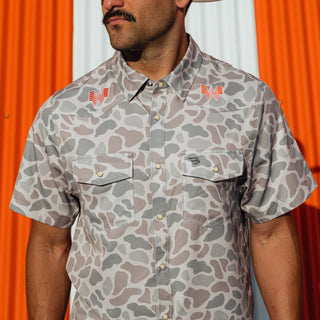 A man wearing a short-sleeve button-up shirt with a Classic Deer Camo pattern and orange embroidered logos on the chest, posed against a background of orange and white drapes.