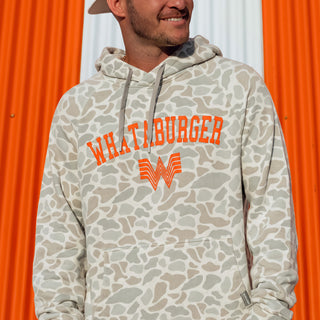 A man wearing a Classic Deer Camo Vintage Washed Hoodie with orange 'Whataburger' text and logo on the chest, standing in front of an orange and white corrugated metal background.