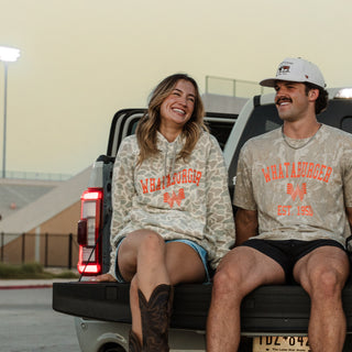 A man and a woman sitting together on the tailgate of a pickup truck at dusk. Both are wearing matching light beige camouflage hoodies and t-shirts with orange 'WHATABURGER'