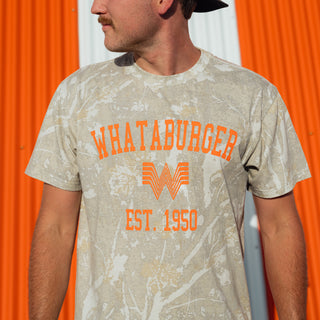 A man wearing a Drifter Camo 1950 Vintage Washed graphic t-shirt with orange text reading 'WHATABURGER' and 'EST. 1950' against an orange and white corrugated metal background.