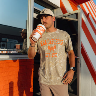A man wearing a Drifter Camo 1950 Vintage Washed Tee with orange 'WHATABURGER EST. 1950' text and logo, standing in front of a restaurant counter and drinking from a branded Whataburger cup.