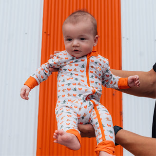 Baby held up in front of orange and white striped building wearing WHATA Mallard Baby Zip Up.