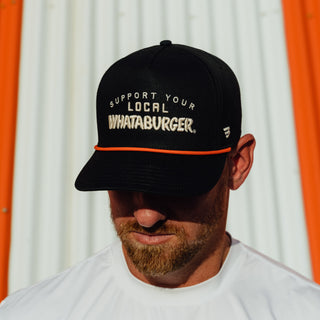 A man wearing a black baseball cap with 'SUPPORT YOUR LOCAL WHATABURGER.' embroidered in white on the front, featuring a thin orange stripe under the brim, set against a white corrugated metal wall with orange side panels.