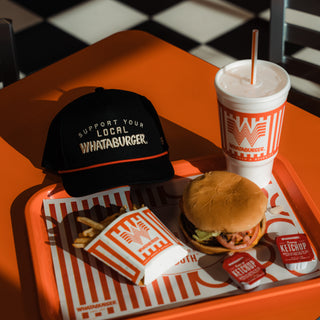 A Whataburger meal set on an orange tray, featuring a burger, french fries, a drink, and a black cap with white embroidered text reading 'SUPPORT YOUR LOCAL WHATABURGER.'.