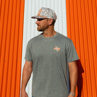 A man wearing a heather gray crewneck t-shirt with a small orange and white graphic on the left chest, paired with a cap, standing in front of a corrugated metal wall with orange and white panels.