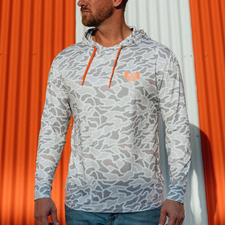 A man wearing a performance hoodie with a white camo pattern, orange drawstrings, and an orange Whataburger  logo, standing in front of an orange and white corrugated metal wall.