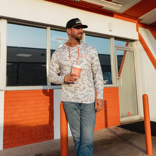 A man wearing a white camo performance hoodie with orange drawstrings and a black cap stands outside a building with orange and white walls, holding an orange and white striped cup.