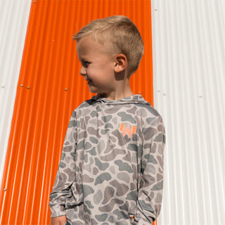 Child smiling and looking to the left wearing Youth WHATA Classic Deer Camo Performance hoodie in front of orange and white striped Whataburger a-frame building.