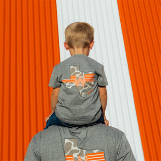Child wearing Youth WHATA Texas Tee sitting on shoulders of dad wearing matching tee in front of orange and white striped Whataburger A-Frame building.