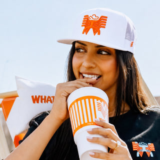 Woman wearing a white hat with an orange logo Whataburger x Hooey, holding a branded cup, with a blurred background.