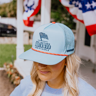 Woman wearing a light blue cap with text and an American flag design, standing outdoors.