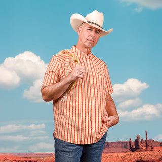 Man in a cowboy hat and striped shirt standing in front of a desert landscape with clear blue sky.