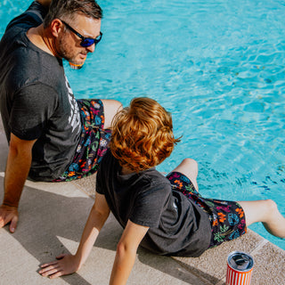 Father and son sitting by a pool, wearing matching Neon Collage Swim Trunks.