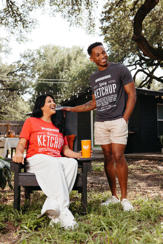 A man and a woman wearing matching Whataburger graphic t-shirts with Ketchup labels printed on the front, posed outdoors in a backyard setting with trees and a grill visible in the background.