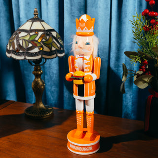 View Whataburger 2024 Nutcracker. Sitting on a wooden table alongside other a holiday lamp and holiday floral arrangement. 