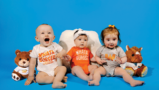 Three babies wearing Whataburger onesies and sitting alongside Whataburger plush