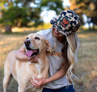 view woman sitting in a field with her dog. Wearing a Whataburger staunch camo hat and a Whataburger channel letter camo shirt.