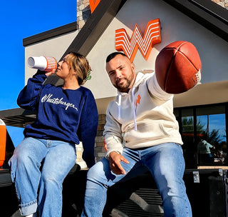 view Woman drinking out of a 30 ounce whataburger yeti while wearing a sportiqe crewneck sweater featuring a whataburger script lettering. The man sitting next to her is extending a football as if to hand it to you, he is wearing the sportiqe hoodie featuring the whataburger flying w logo.